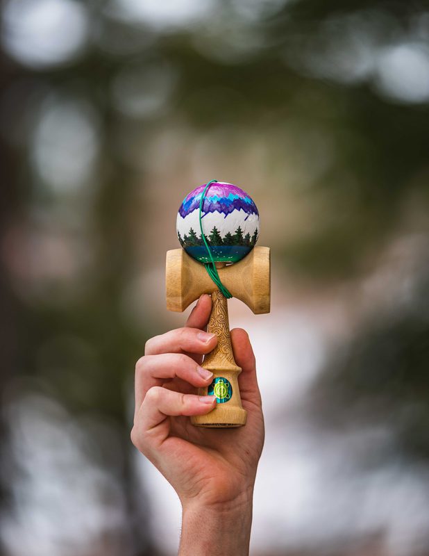 Hand holding a mountain-painted kendama toy against a forest backdrop